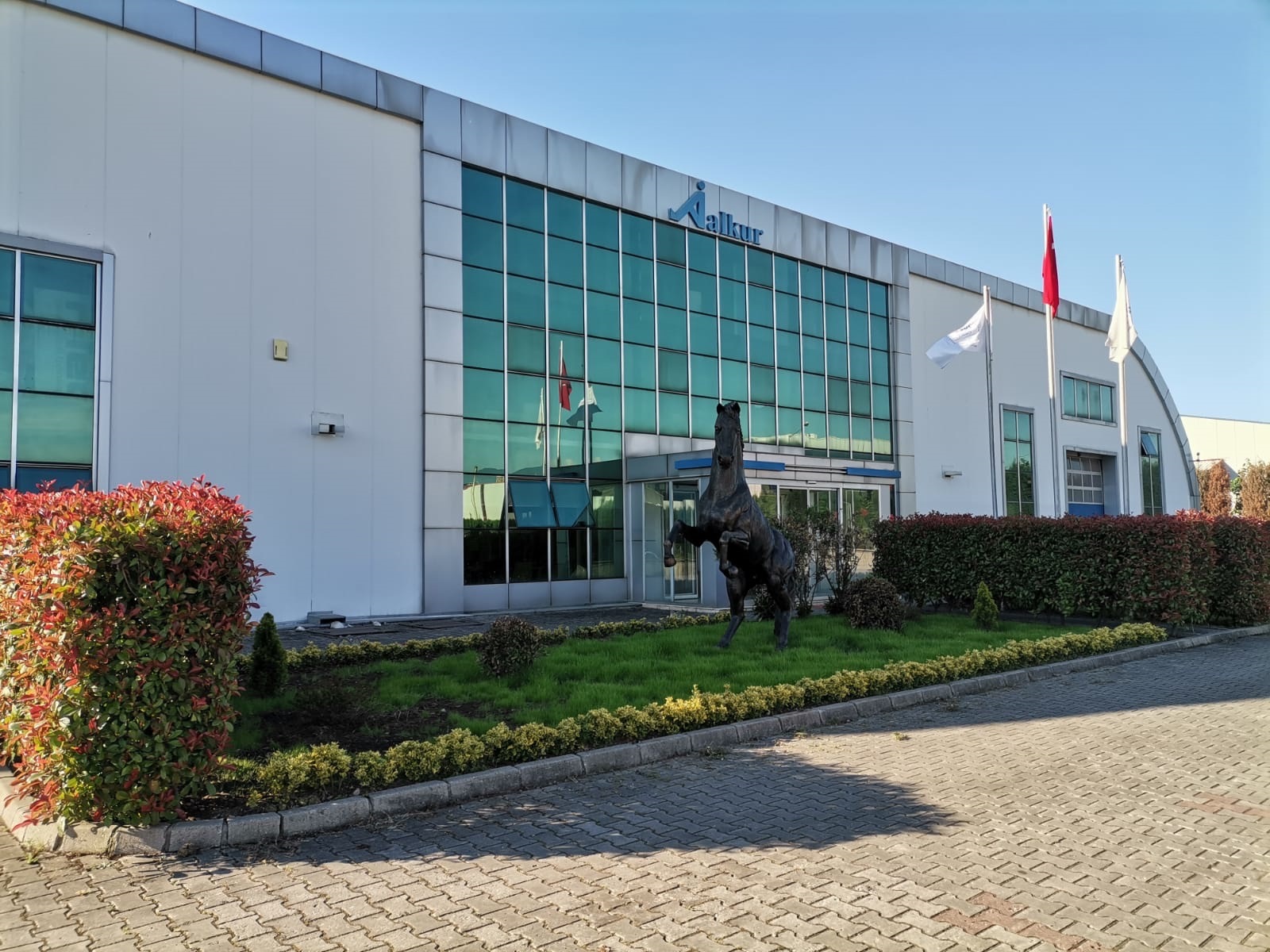 Alkur fabrika dış cephesi — cam cephe, logo ve giriş bahçesi.
